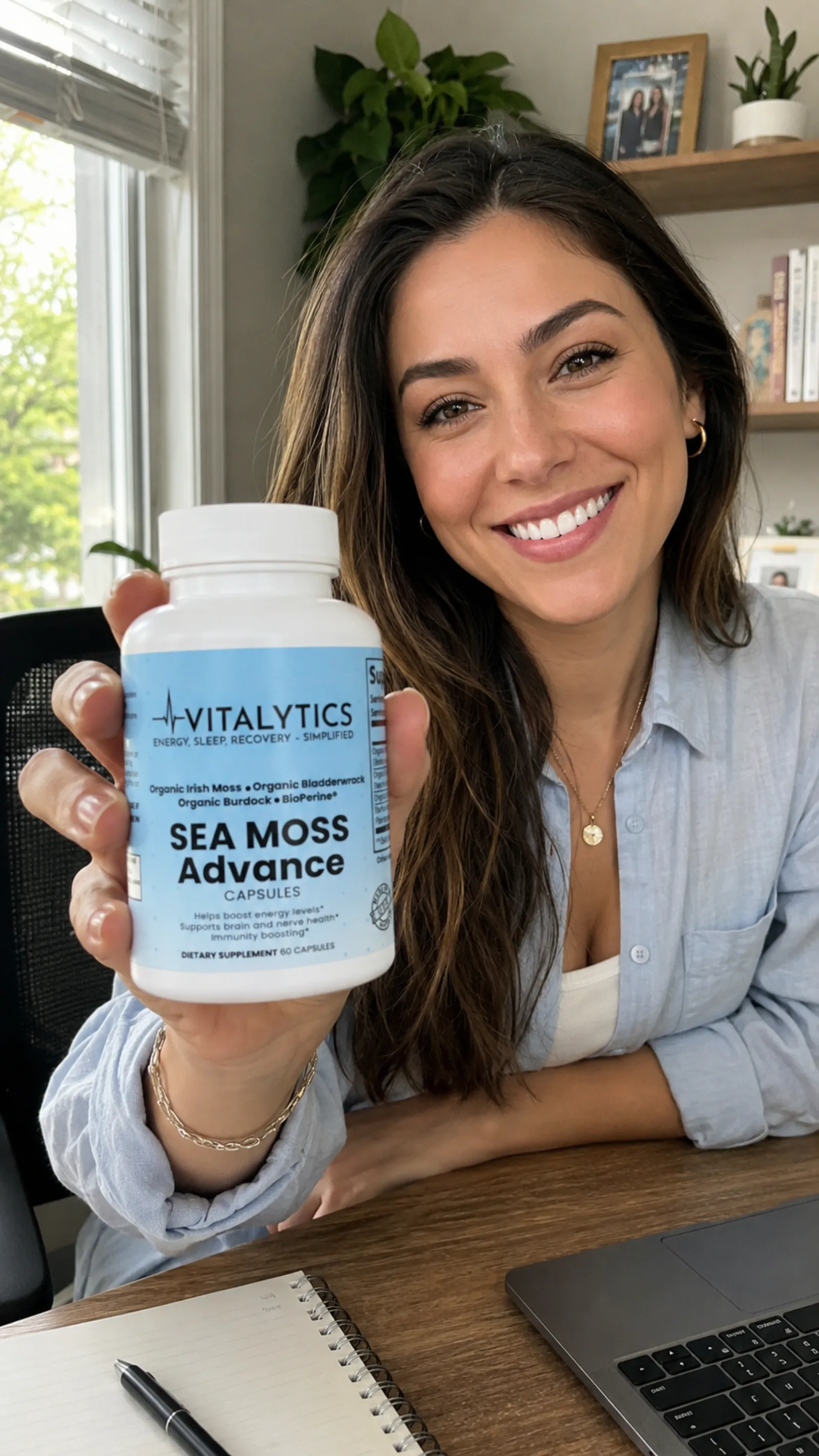 Woman at a desk smiling and holding Vitalytics Sea Moss Advance beside a notebook and laptop.