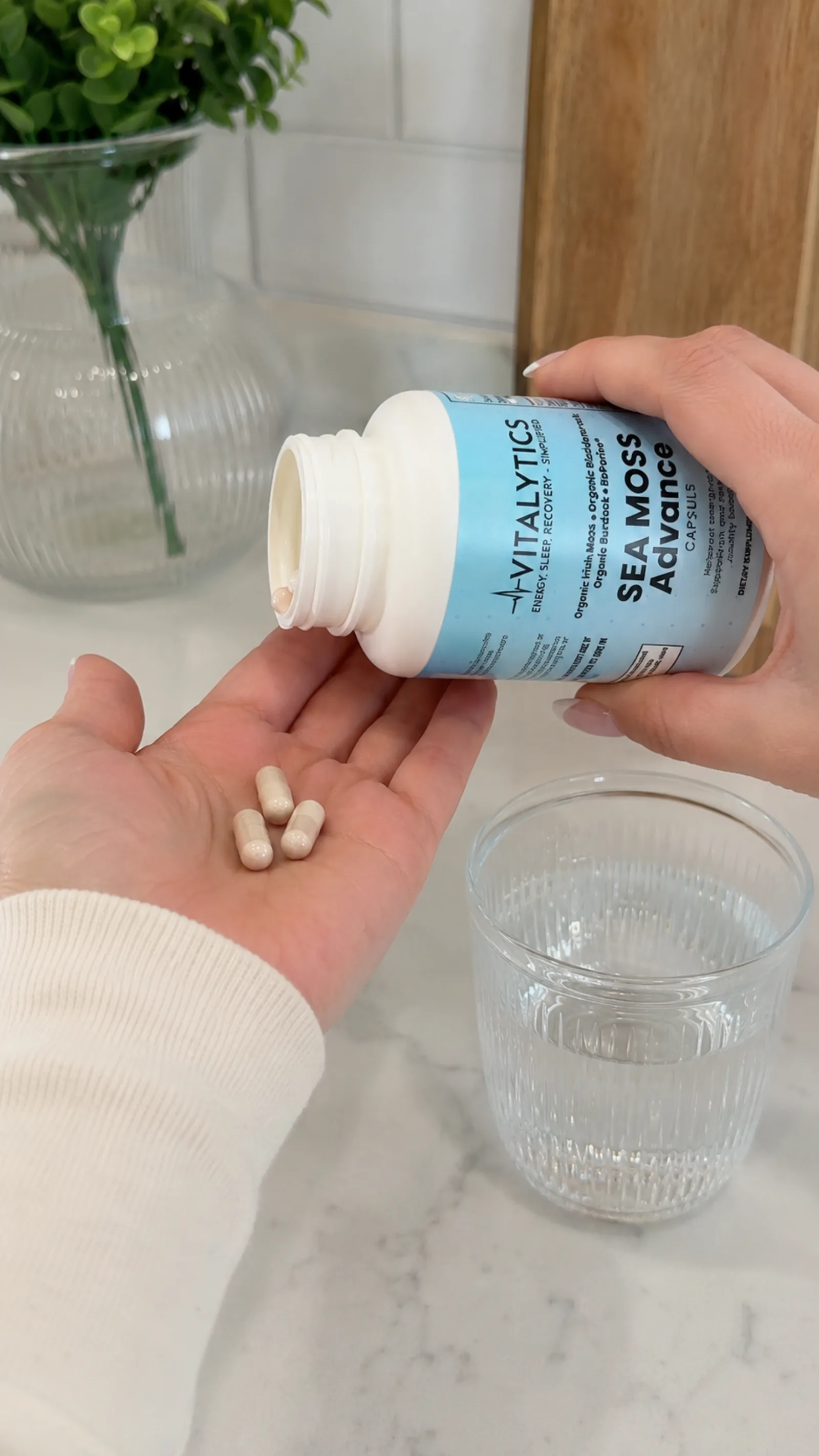 Close-up of Vitalytics Sea Moss Advance capsules being poured into a hand beside a glass of water.
